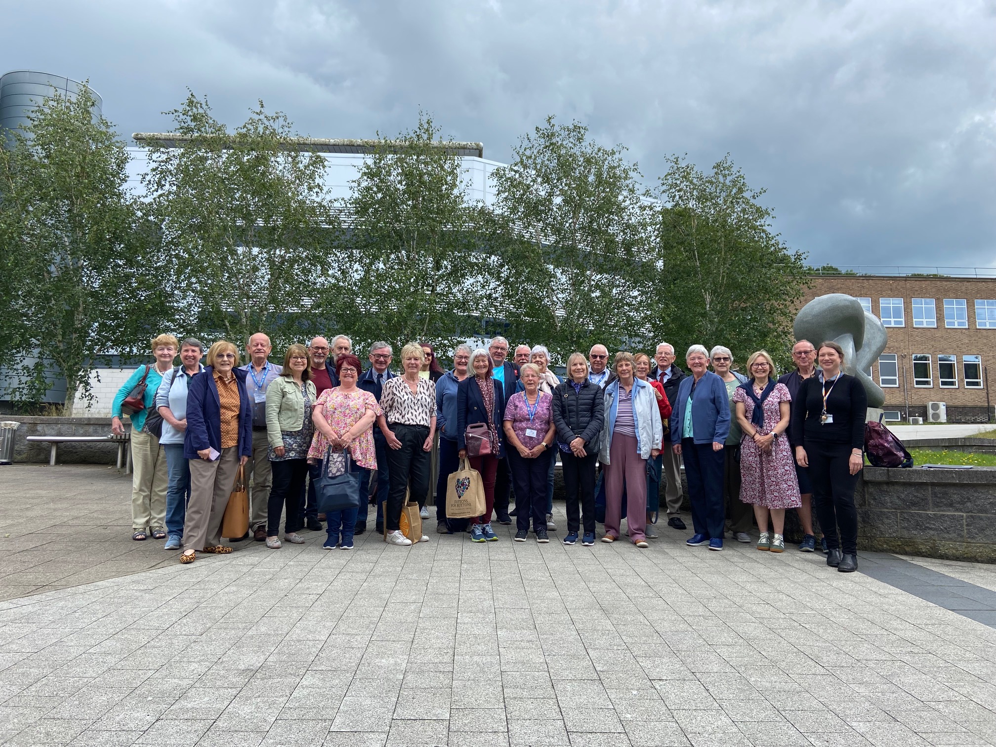 Group photo of smiling members of Northumbria Region U3A, standing near campus buildings and trees under a cloudy sky.