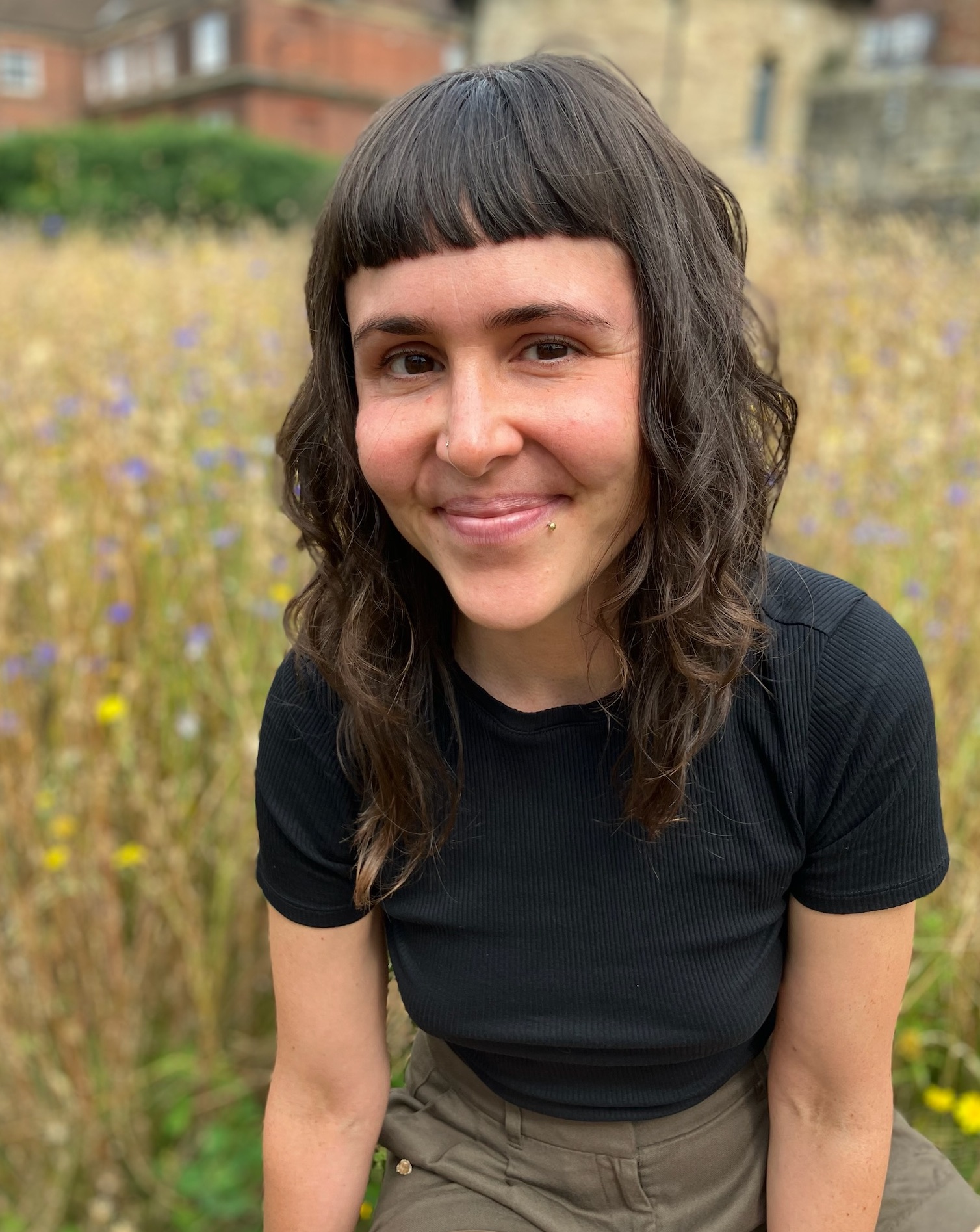 Dr Kelly Blevins is sitting and smiling in a flowery garden. In the blurry distance are some buildings.