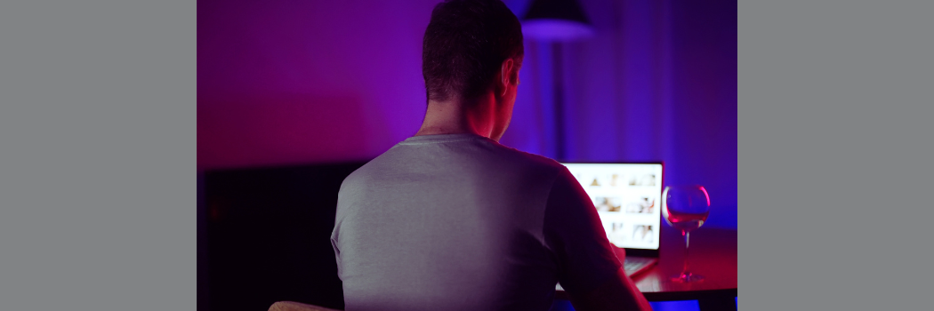 A man sits in the dark in front of a computer with a glass of wine