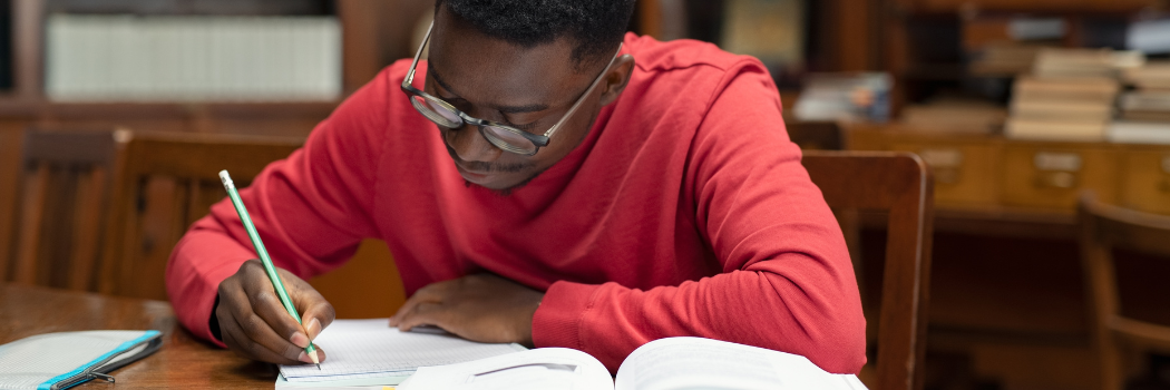 Young black man with glasses taking notes from book while sitting in the library.