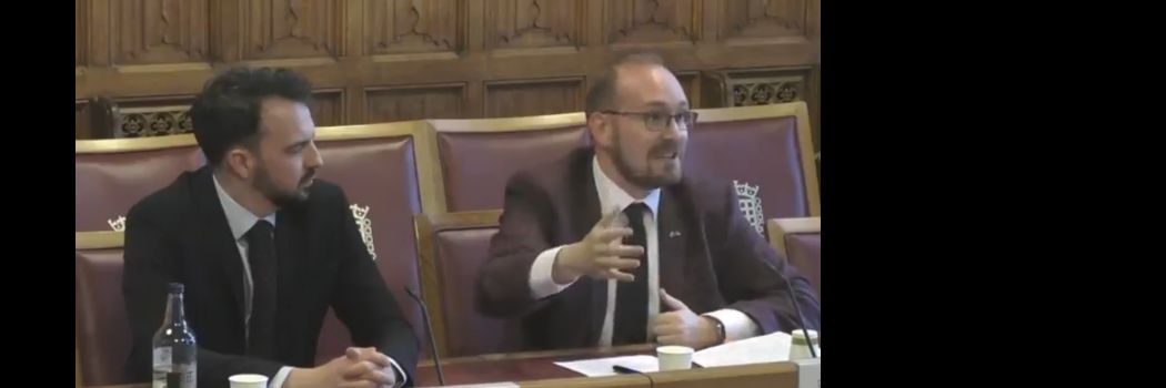 Two people sitting at a desk with papers, addressing an audience off-camera