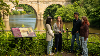 A group of 4 students stand by the river in front of a bridge