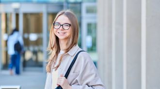 Student with blonde hair and glasses smiling at camera outside the business school