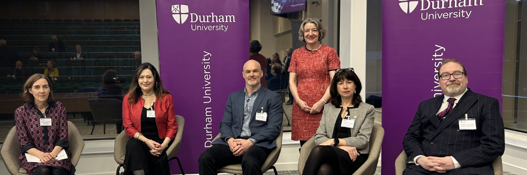 A mixed group of men and women sitting down. A women in a red dress stands behind them in between two purple 'Durham University' banners