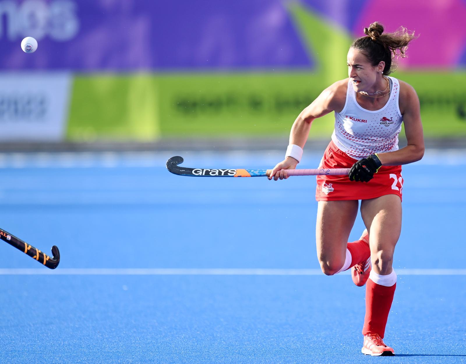 A female hockey player wearing England kit running towards the camera on a blue astroturf