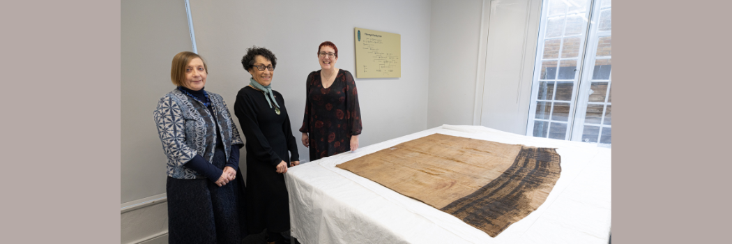 Three women standing next to an ancient cloak, laid out on a table