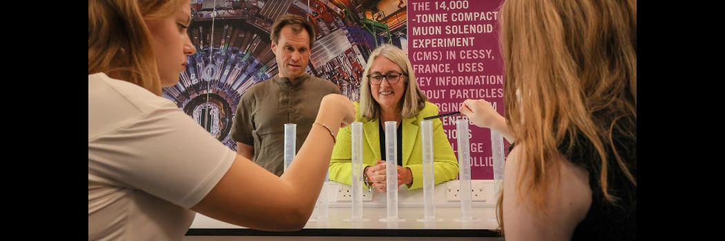 Two women and a man standing behind a science desk, watching two young women point tuning forks at plastic tubes on a table