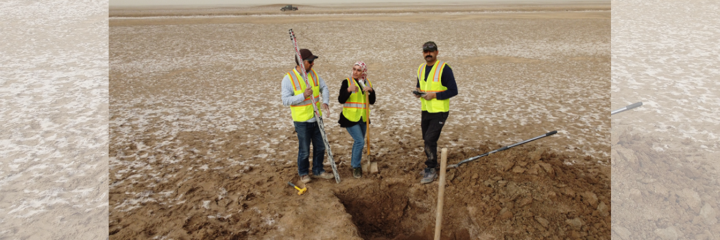 Two male and one female archaeologists stand by a deep trench in a desert landscape