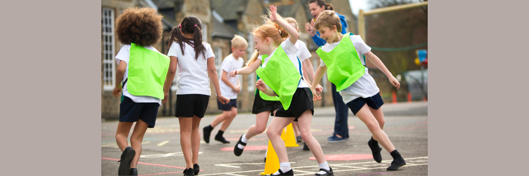 A mixed group of primary school children running in a circle in a playground with a teacher in the background.