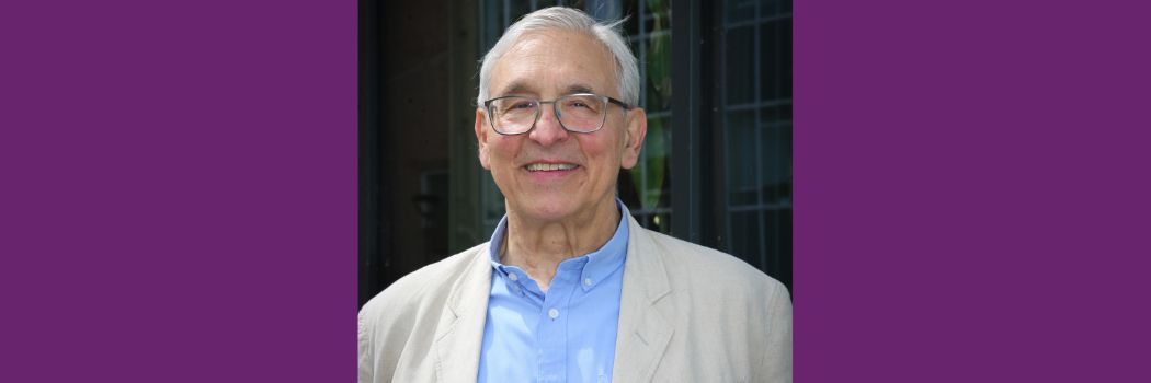 A man with white hair and glasses, wearing a cream linen jacket and blue shirt smiles at the camera
