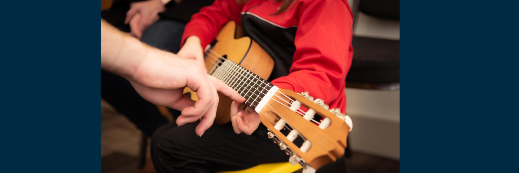 Close up of a child holding a guitar as an adult points to where he should put his fingers