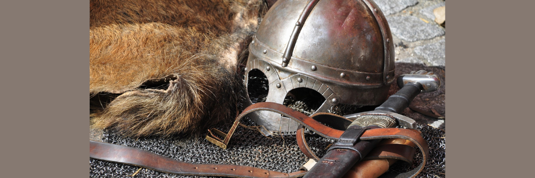 A close up of a medieval metal helmet and sword lay on the floor next to a brown fur cape.