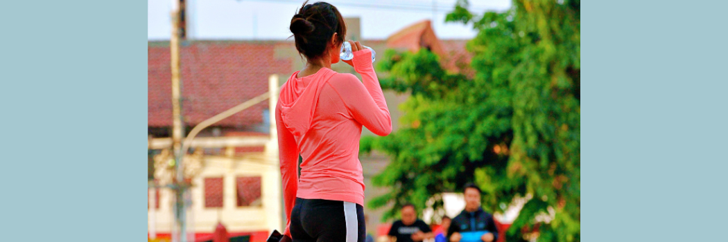 The back of a woman outdoors wearing a bright pink top and black leggings drinking from a water bottle.
