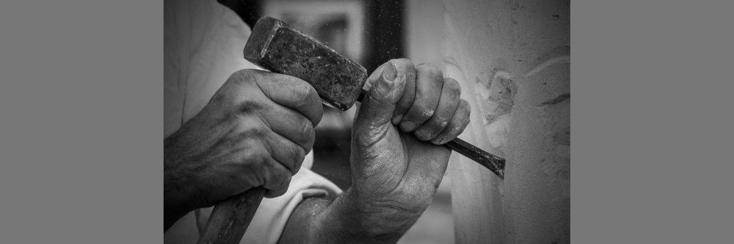 A black and white close up of the hands of a stonemason holding a chisel and hammer against a wall