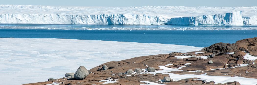 A snowy, rocky foreground with a white glacier, blue sea and blue sky.