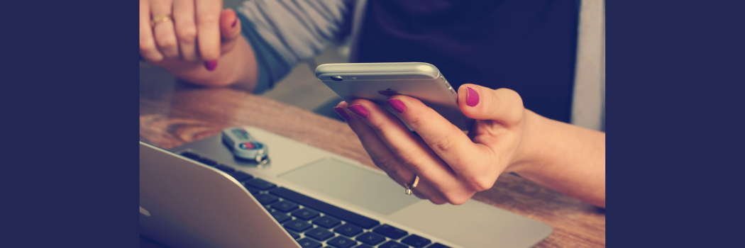 A close-up of a woman's hand holding a silver mobile phone above a laptop computer on a desk.