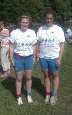 Two girls with white 2013 Durham regatta t-shirts on smiling at the camera, standing on the banks of the River Wear