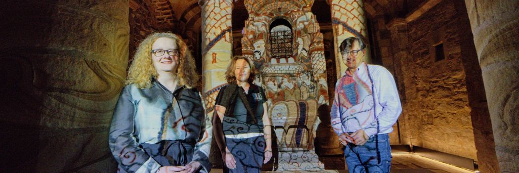 Three people smiling at the camera in an old chapel with sandstone pillars. A colourful projection covers them and the wall behind them. The projection is reminiscent of The Bayeux Tapestry.