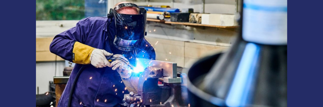 A man wearing blue overalls, a welder's helmet and visor using a tool.