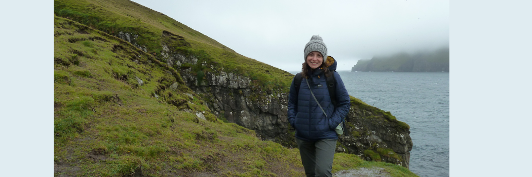 A female in a coat and grey bobble hat standing on the side of a grassy hill with mountains in the background