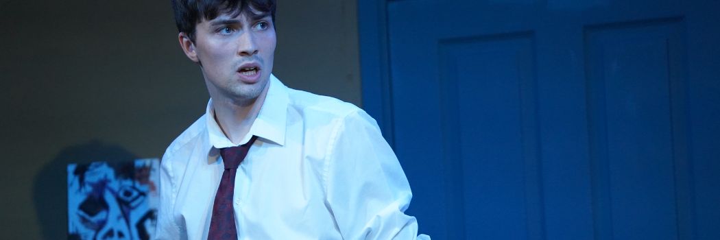 A young man wearing a white shirt and dark tie on a theatre stage