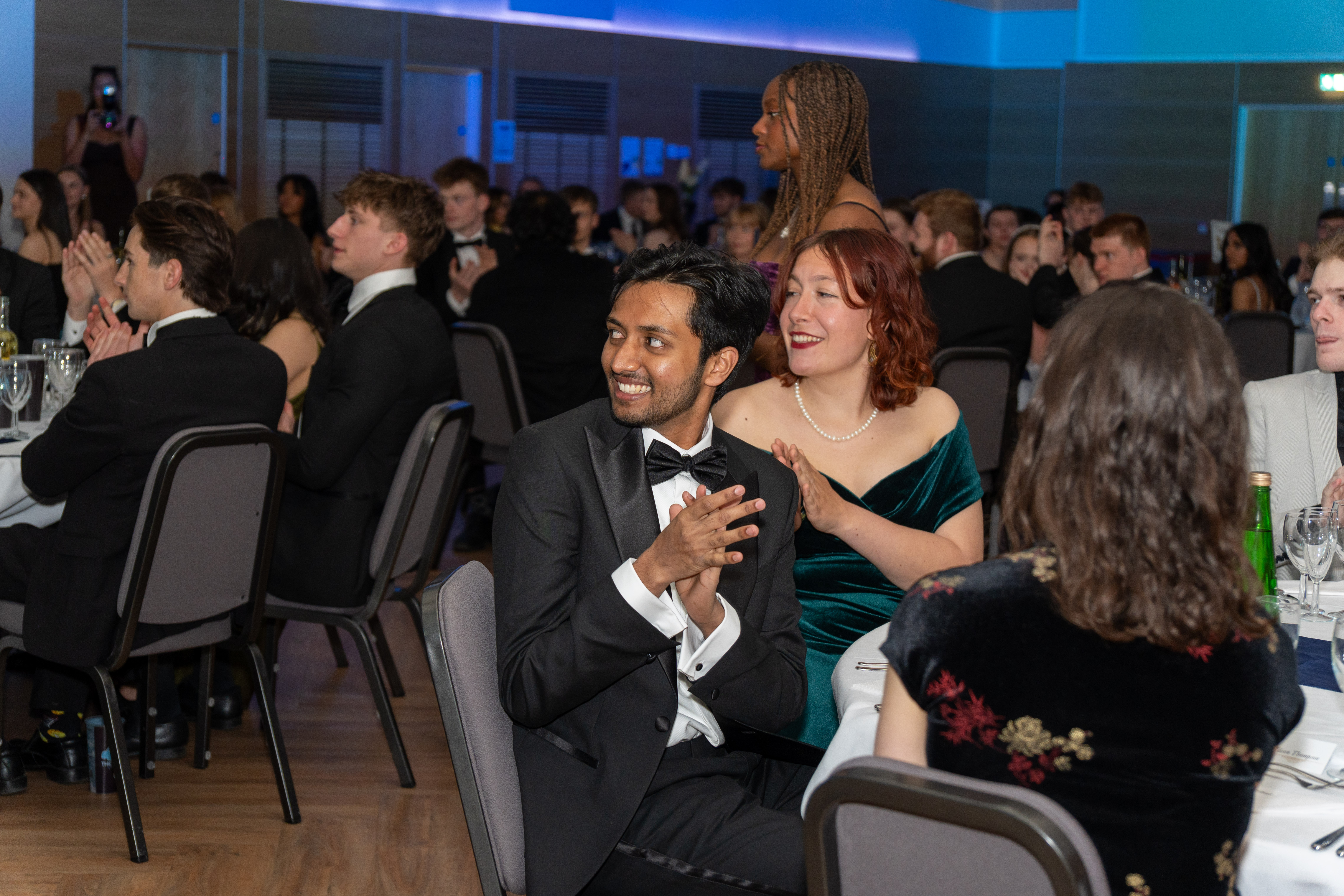 Guests in formalwear applaud during the annual awards ceremony at John Snow College. Seated at round tables, they watch the event unfold in a warmly lit room with a blue ambient glow, creating a celebratory atmosphere.