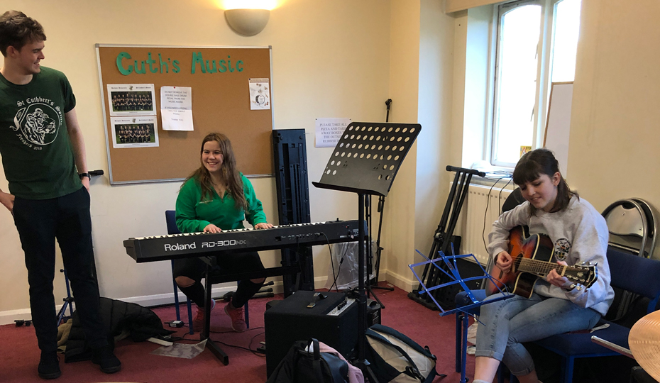 Students in the music practice room with a drum set in the foreground. One student playing keyboards, one playing the guitar, and the other singing.