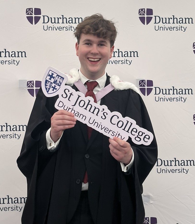 a student in academic dress smiles at the camera holding a St John's sign