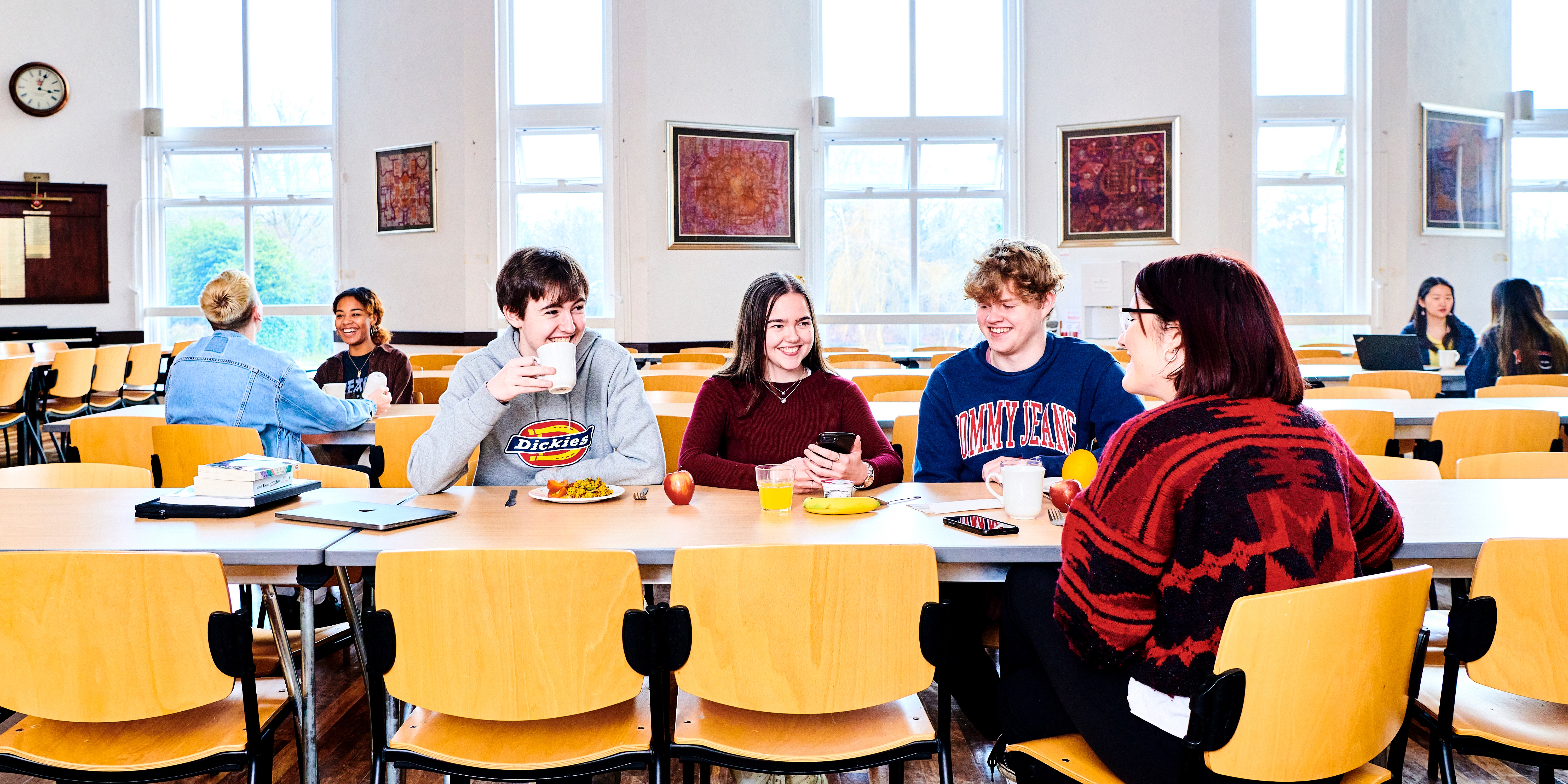 Male and female students sit at long tables, eating and socialising. The dining hall is brightly lit, with high ceiling and large windowstbc