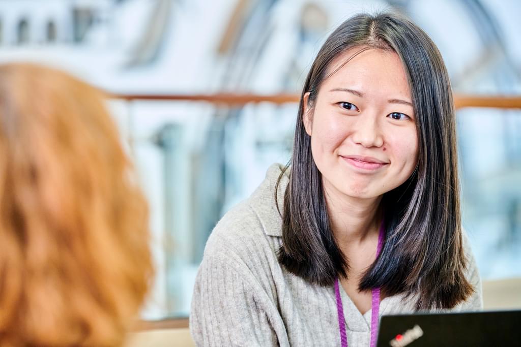 Woman with long black hair sits in a brightly lit space smiling at colleagues. She is a Durham Uni alumna (Masters of Science) and works as a co-ordinator in the International Office