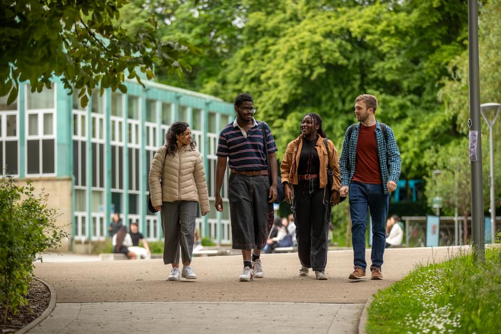 Four students walk towards the viewer. They are chatting as they walk. In the background there is a zinc green building and lots of trees in full leaf.