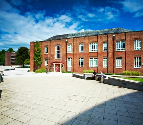 Photo on a sunny day of a red brick building, Durham University's Dawson Building, with a pale grey paved surface around it and some cultivated pockets of grass with nearby benches. The sky above the building is a bright blue.