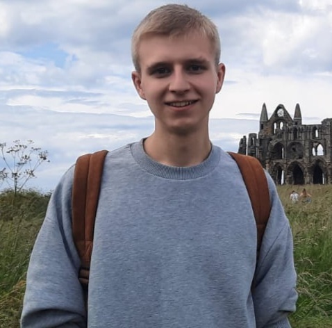 Head and upper torso photo of staff member Max Storey. Max stands facing the camera, in front of a grassy field with building ruins in the far background. Max wears a bluish-grey sweater and a backpack..