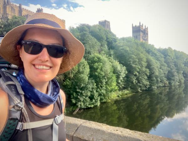 Megan Olshefski, dressed in hiking gear, a hat and sunglasses, smiling at the camera from a bridge over the river with Durham Cathedral and trees in the background.
