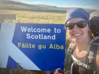 Megan Olshefski smiles at the camera next to an England-Scotland border sign that says 'Welcome to Scotland' and 'Fàilte gu Alba'. In the background are fields bright in the sunlight.