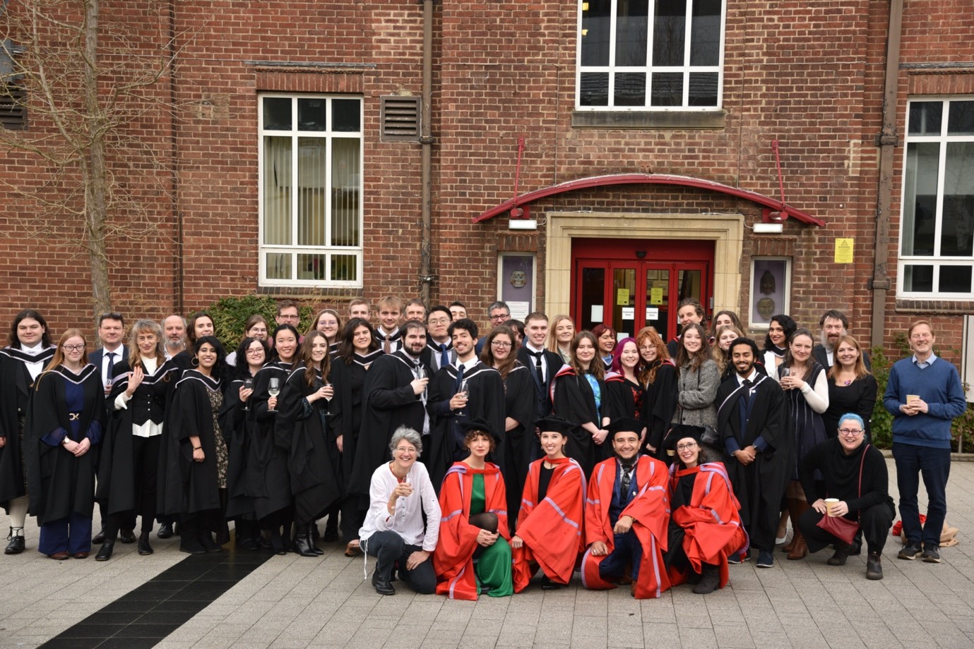 A large group photo in front of the red-brick Dawson Building, with most of the group wearing graduation gowns.