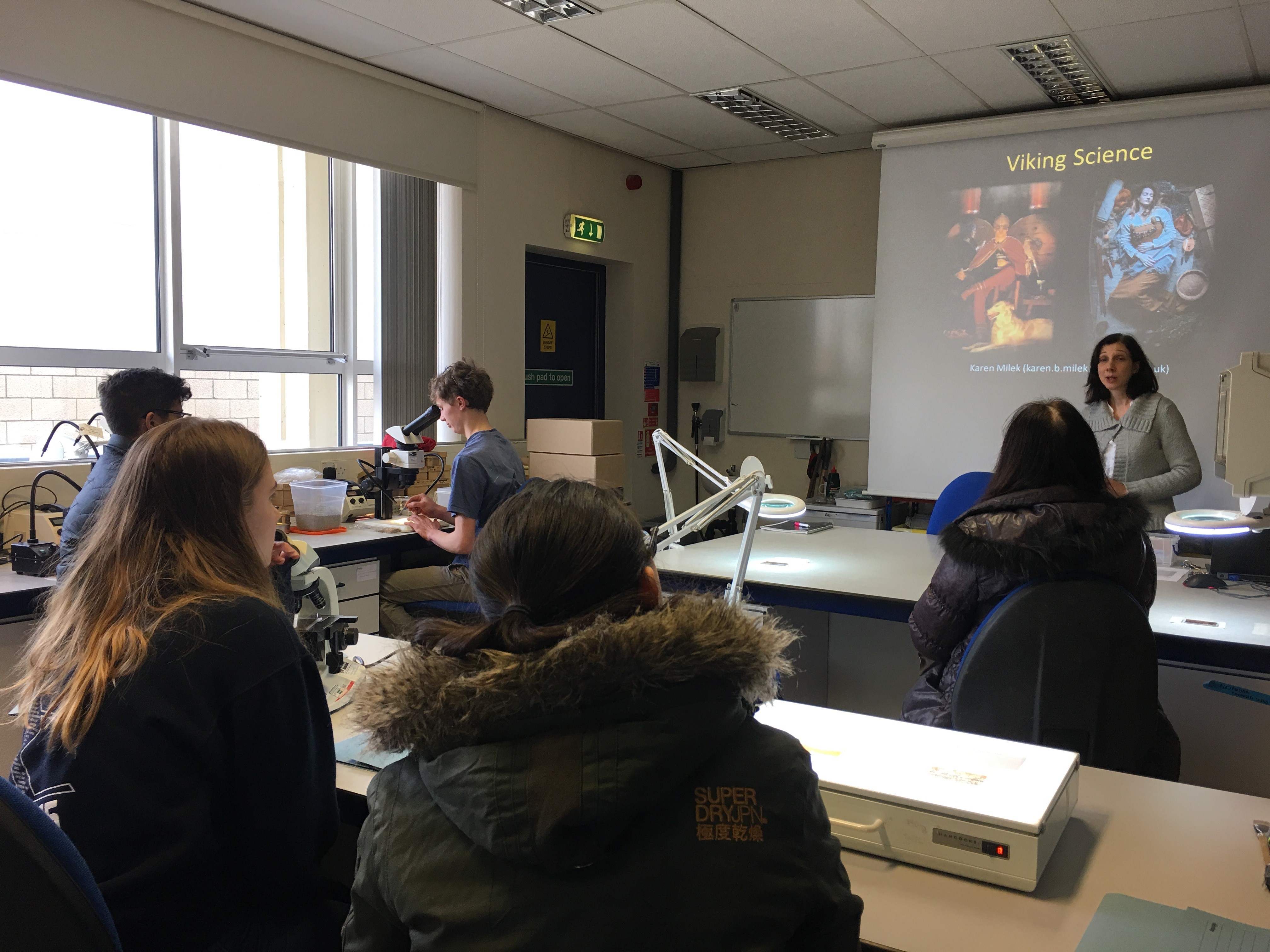 Students sit in an Archaeology laboratory workspace. They are being taught by a lecturer at the front of the lab who stands in front of a projected presentation titled Viking Science. Around the lab are microscopes, magnifying lamps and other equipment.