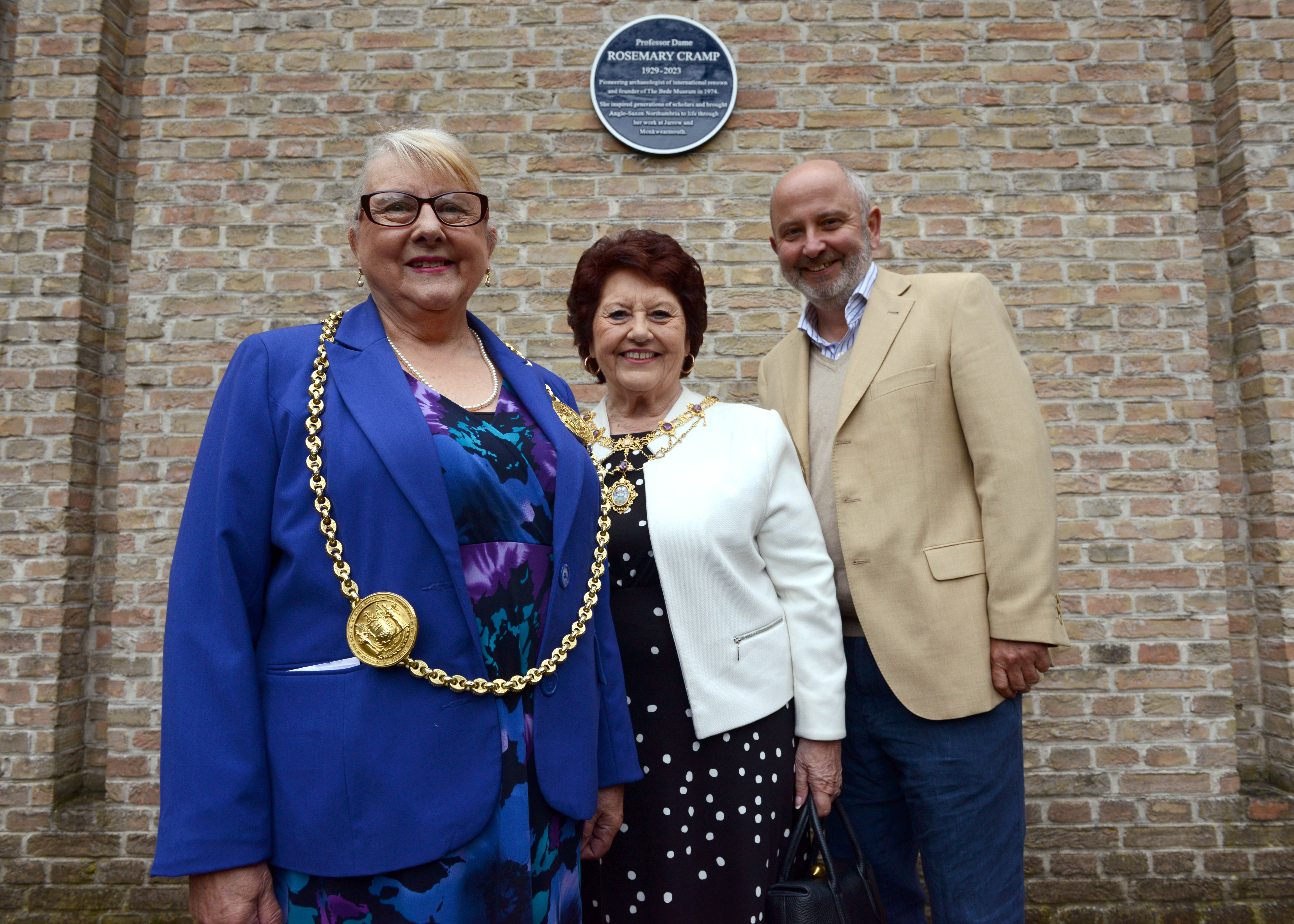 Three smartly dressed and smiling people, Councillor Fay Cunningham, Mayoress Stella Matthewson and Professor Chris Gerrard, stand in front of a wall that features a blue plaque honouring Professor Dame Rosemary Cramp, 1929 - 2023.