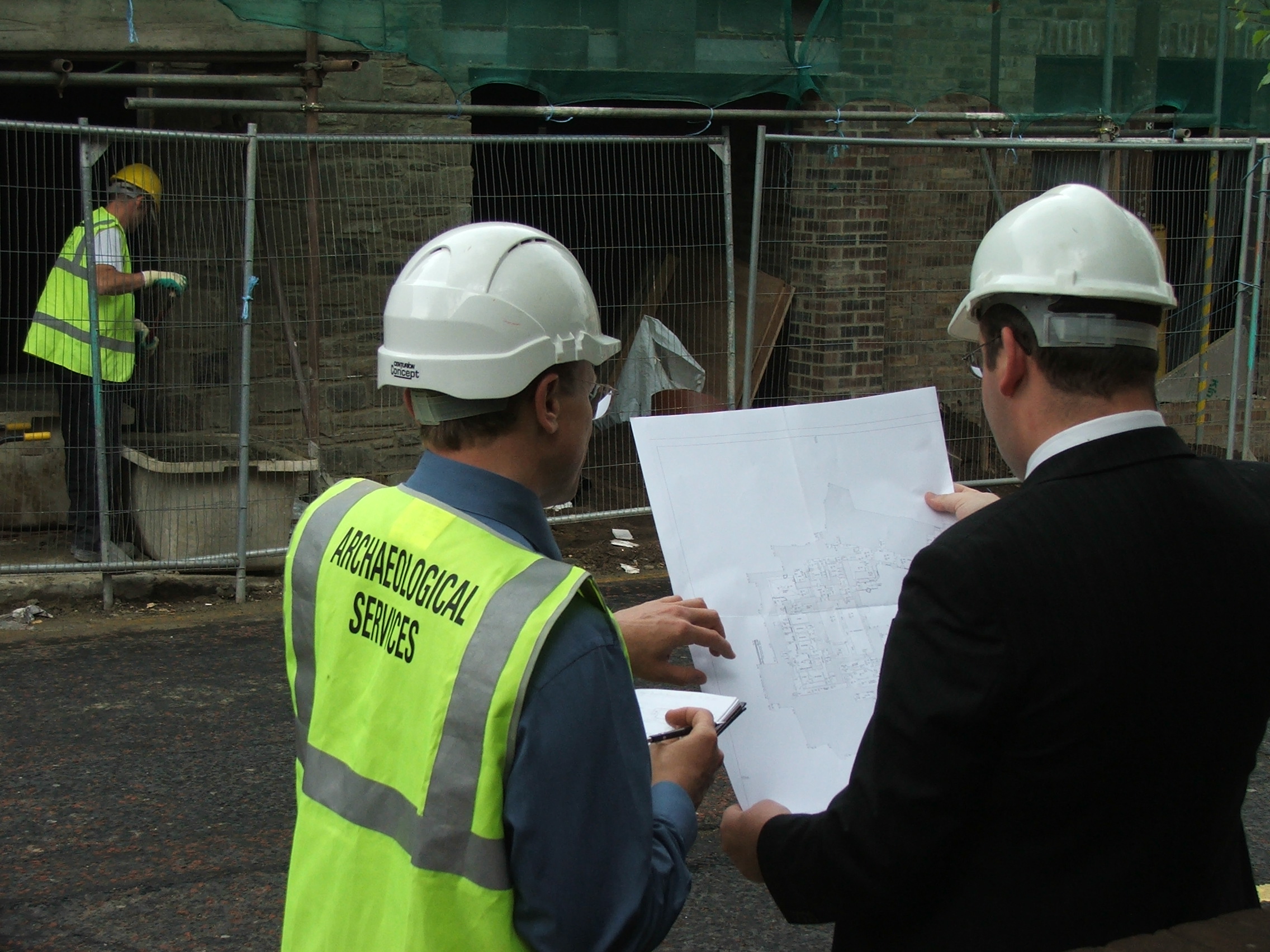 Tan archaeologist and a man in a suit looking at plans in front of building work on a historic building