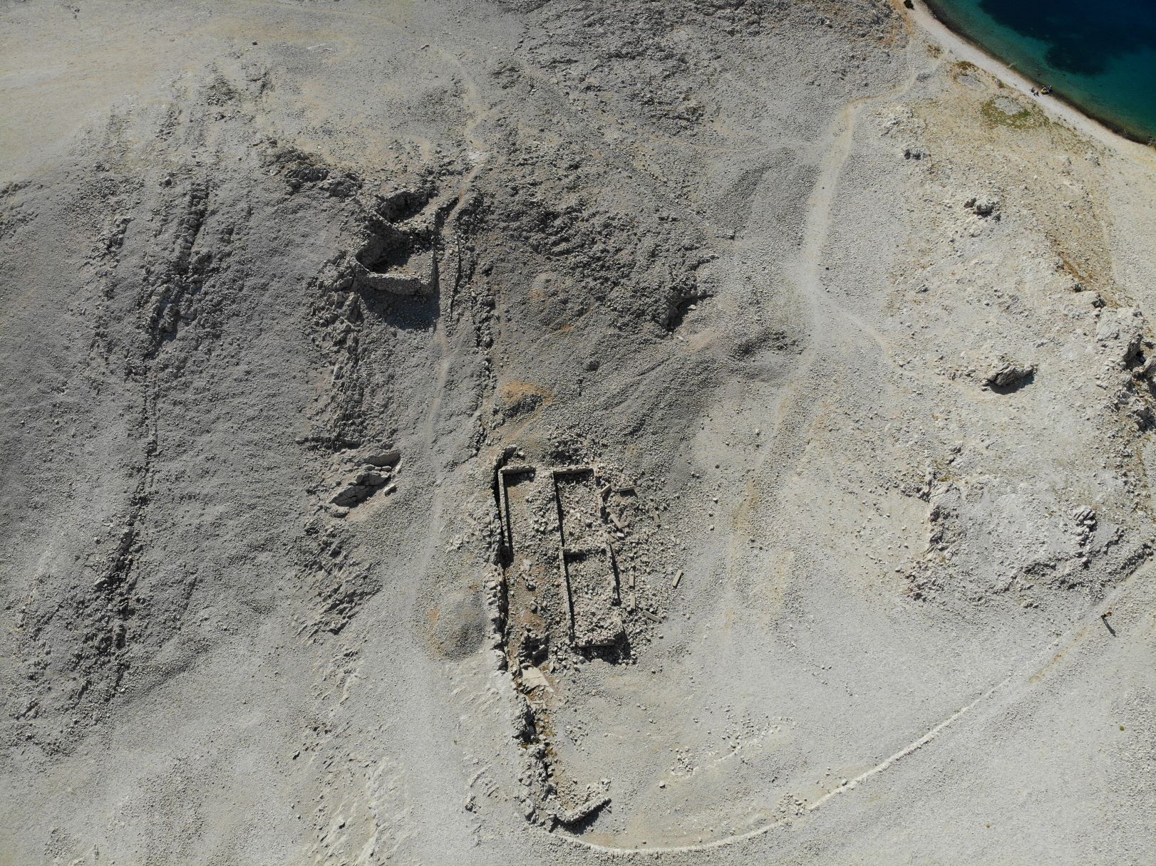 An aerial view of structural remains in rocky terrain. In the top right corner, a blue lake is visible