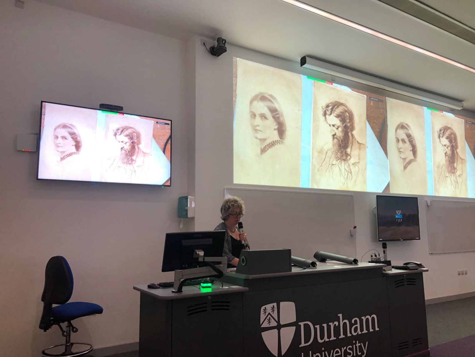 Debbie Challis speaks into a microphone behind a Durham University lecture podium, next to several projector screens, presenting to an out-of-frame lecture theatre audience.