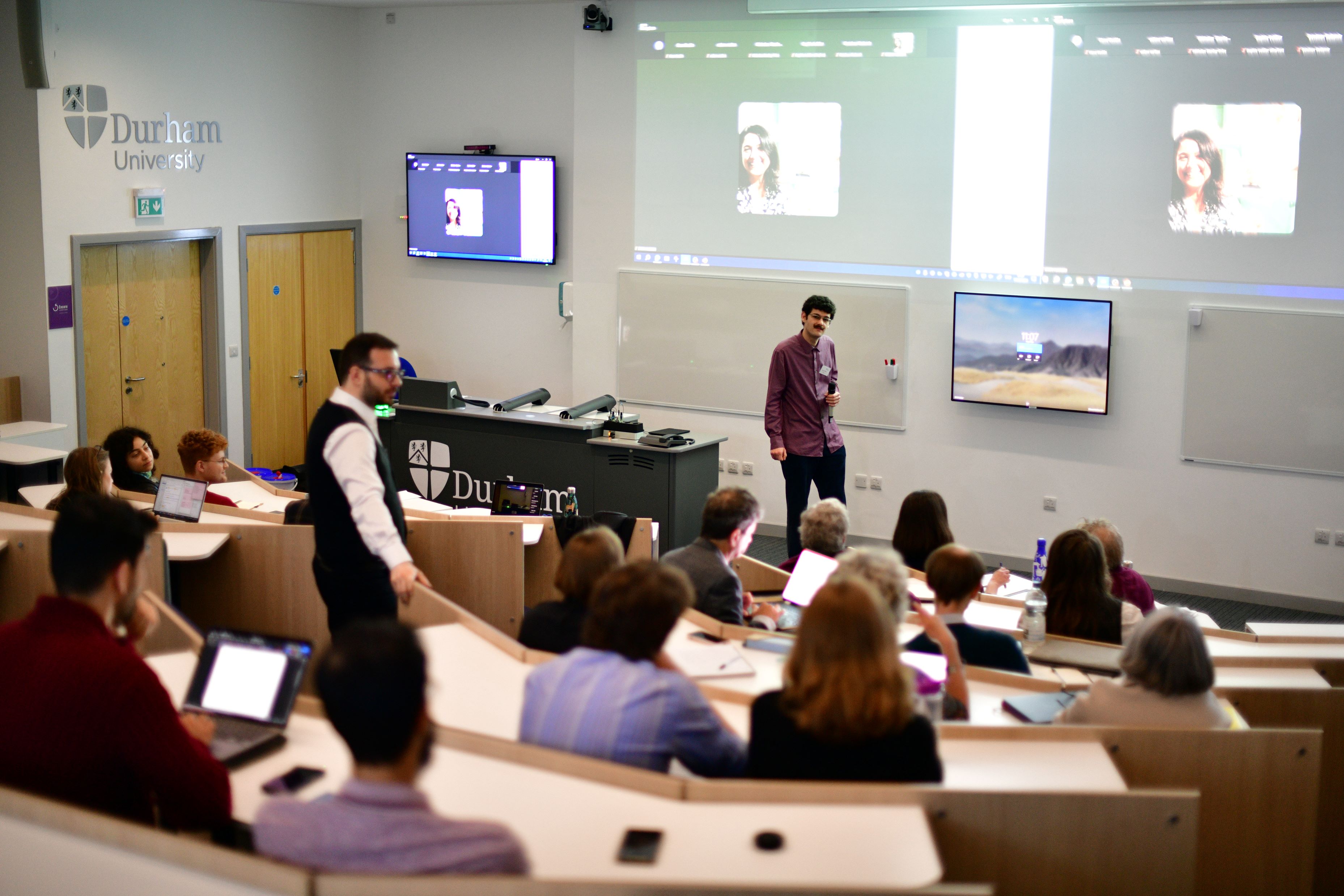View from a back of a full lecture theatre with tiered seating. One conference organiser stands on the central steps halfway up the theatre, another stands in the front of the lecture theatre. People are engaging in discussion and many have their laptops open or paper out on the tables to take notes. There are screens at the front of the lecture theatre with people attending the event online.