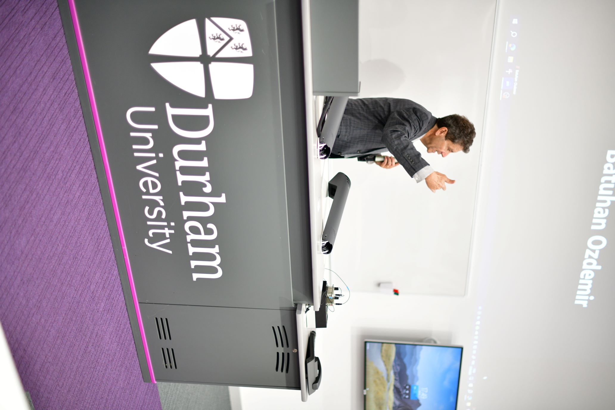 A man in a grey suit, standing behind a Durham University lecture podium, holding a microphone and presenting to an out-of-frame lecture theatre.