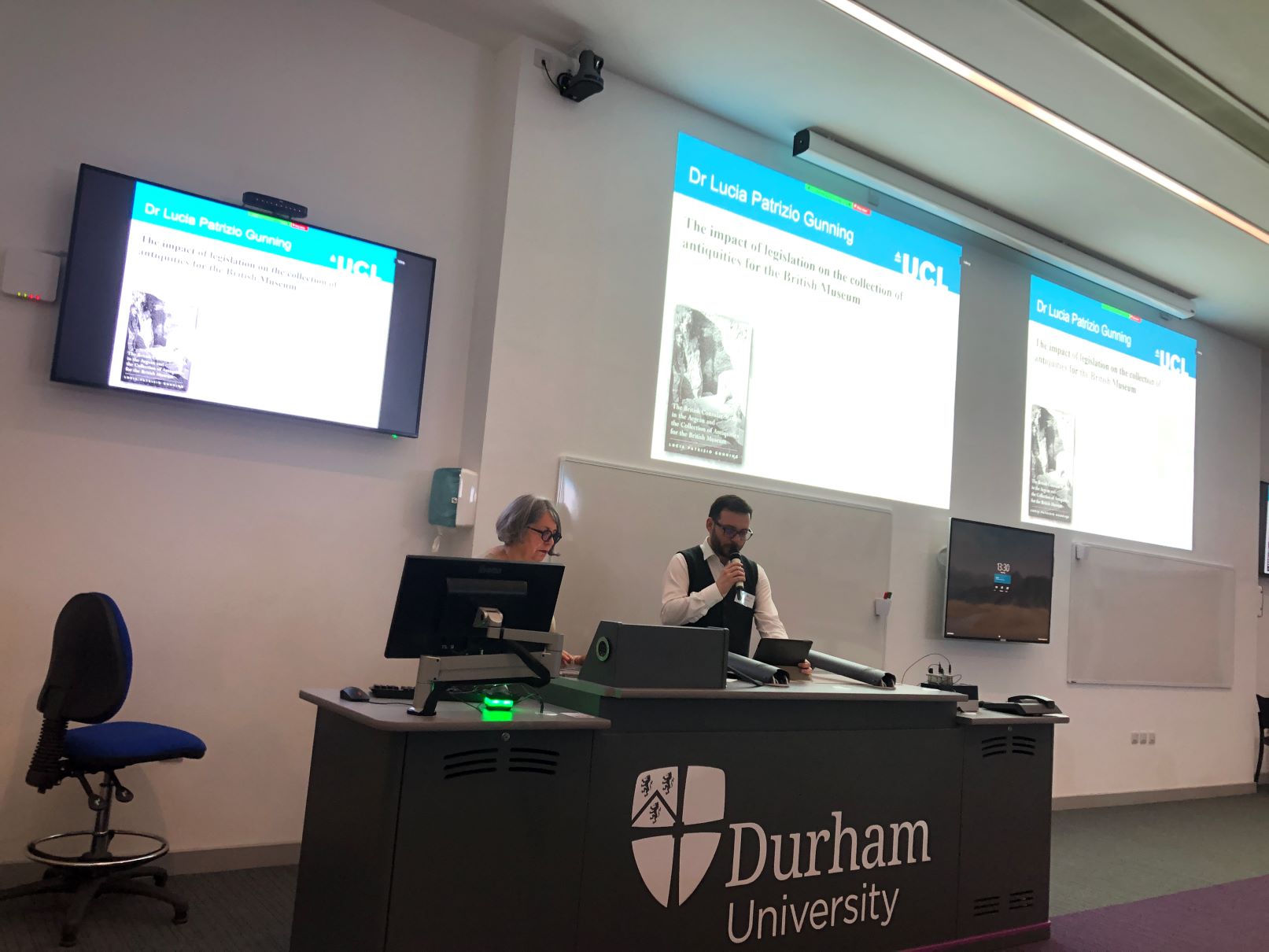 Lucia Patrizio Gunning and Batu Ozdemir stand behind a Durham University lecture podium, presenting to an out-of-frame lecture theatre audience.