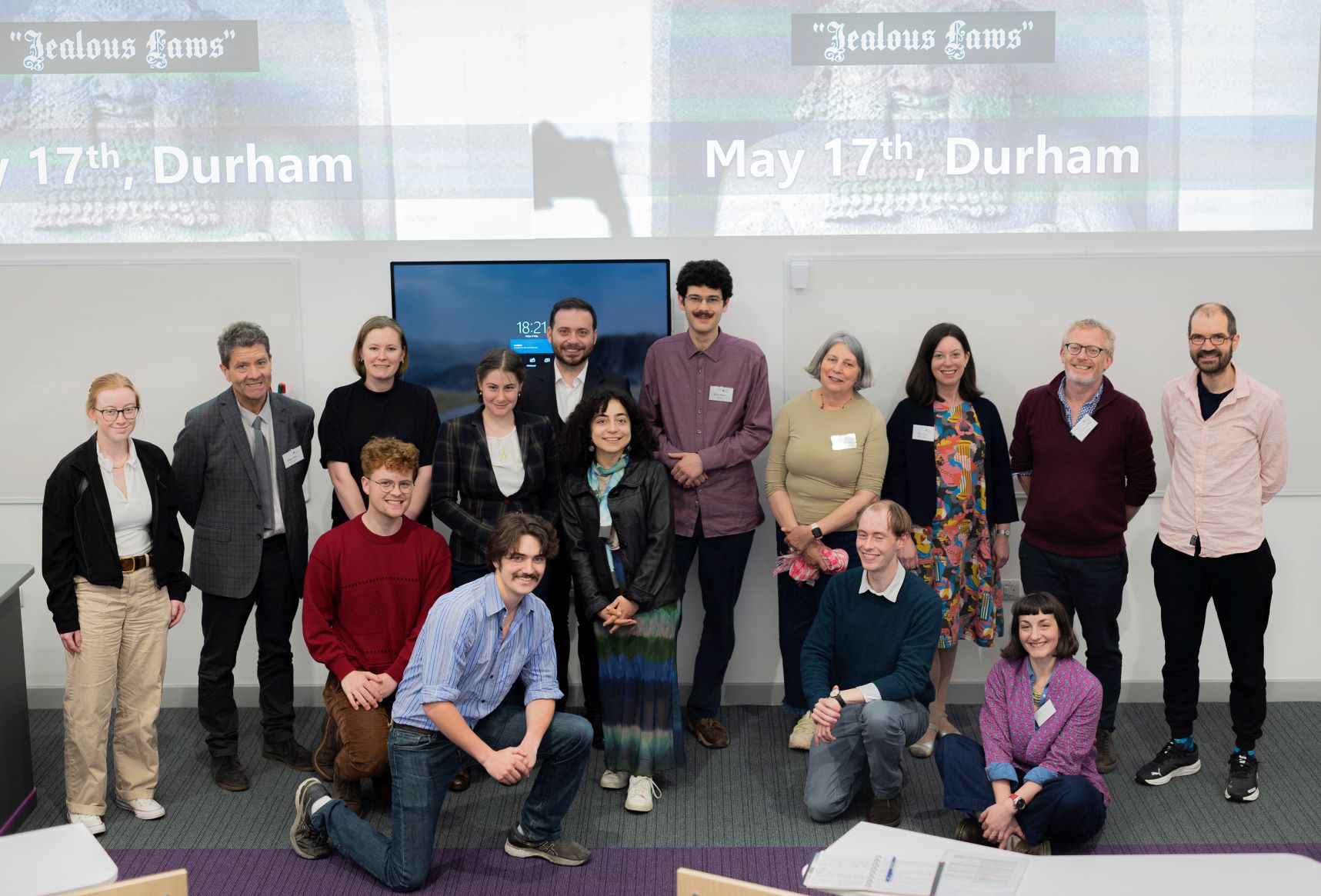 Group of smiling people gathered at the front of a lecture theatre. Behind them are screens with text reading 