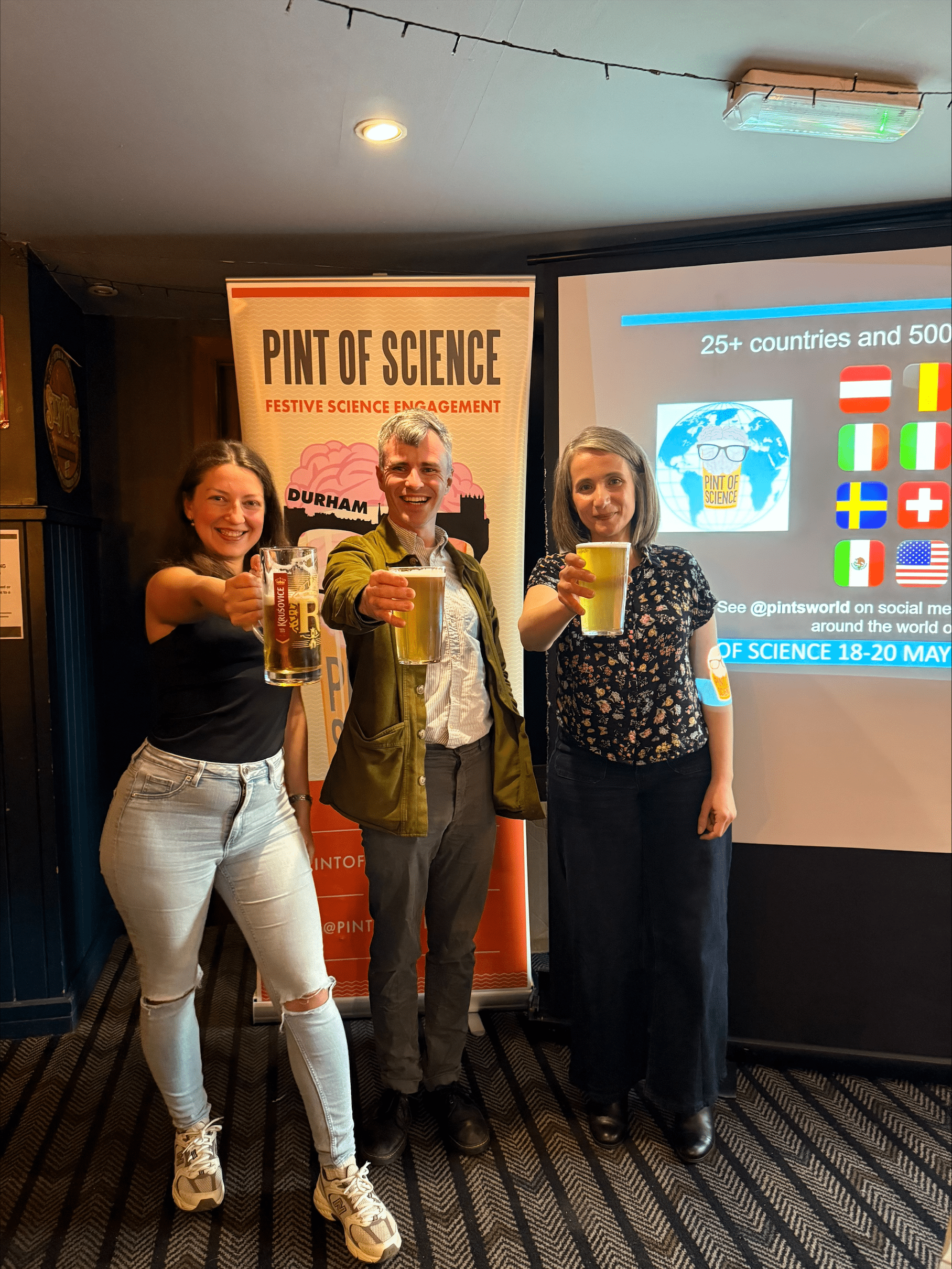 Three people standing in front of a Pint of Science sign