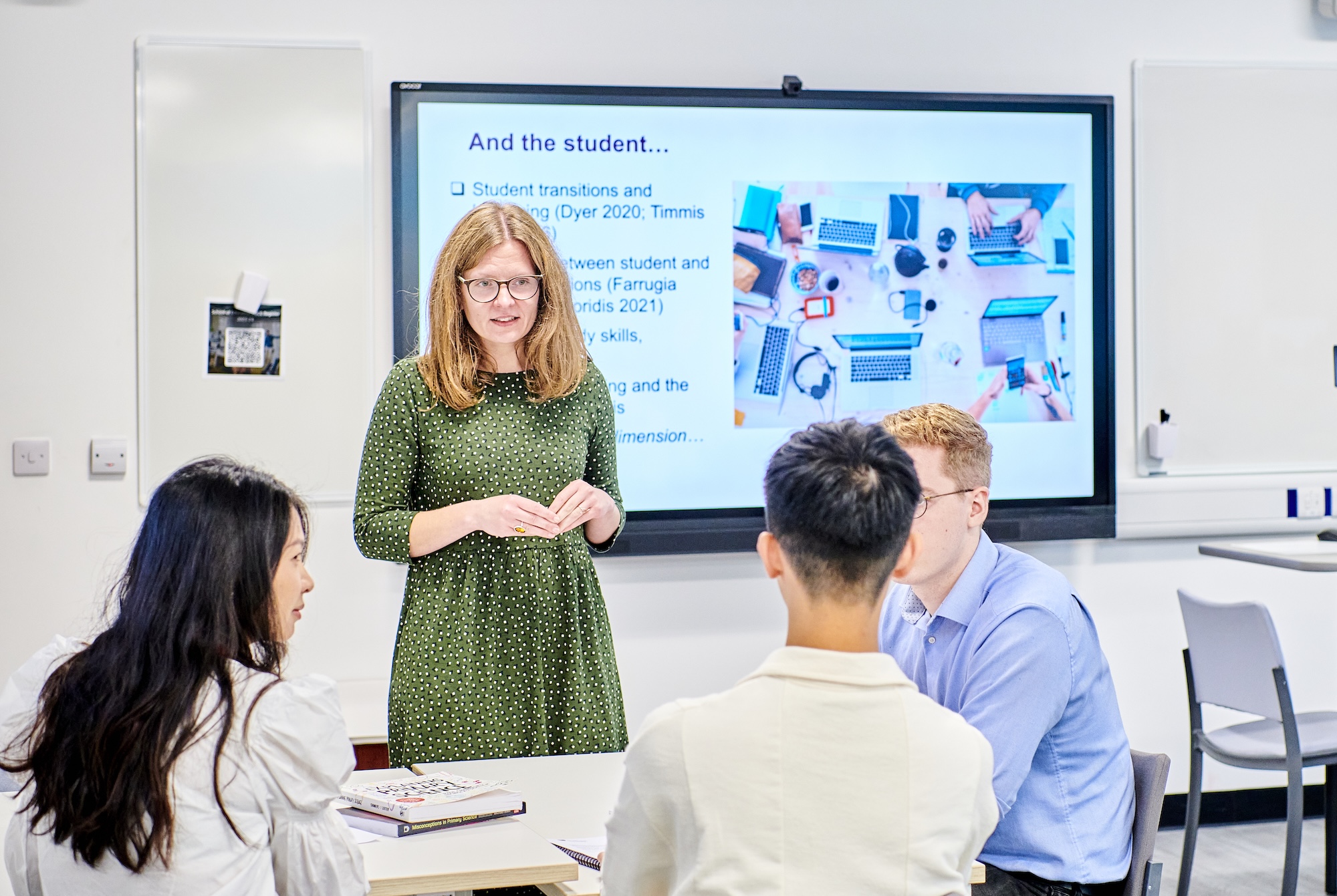A female professor, standing in a green dress, talks to a group of seated students in a seminar room.