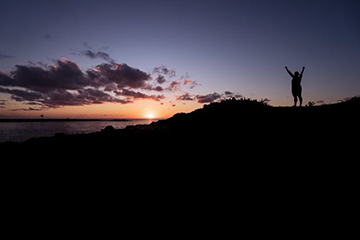 Figure with raised arms looking out at a sunset of the ocean