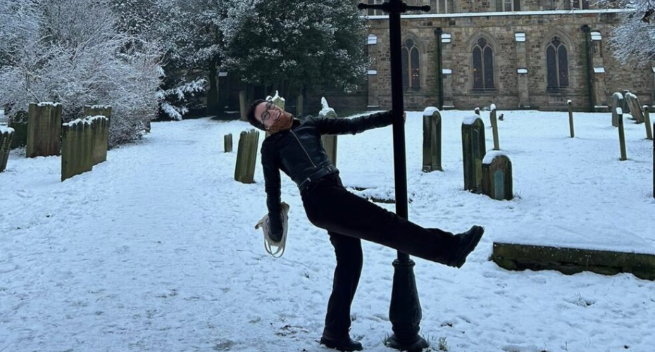 A female student hanging onto a lamp post in snowy weather with a church and gravestones in the background