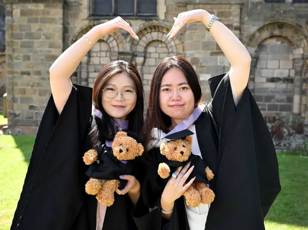 Two students in graduation robes, each holding graduation teddies and using their arms to make a heart shape together.